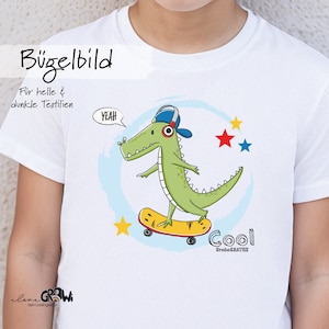 Könnte beinhalten: Ein weißes T-Shirt mit einem grünen Alligator, der einen blauen Hut trägt und Skateboard fährt. Der Alligator sagt "Yeah!" in einer Sprechblase. Der Text "Cool KrokoSkater" befindet sich unter dem Alligator. Der Text "Bügelbild" befindet sich oben auf dem Shirt. Der Text "Für helle & dunkle Textilien" befindet sich unter dem Text "Bügelbild".