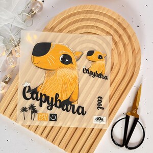 Könnte beinhalten: Ein transparentes Blatt mit einer gelben und schwarzen Cartoon-Capybara-Illustration. Das Capybara ist mit "Capybara" und "Super Cute" beschriftet. Das Blatt enthält auch eine kleinere Capybara-Illustration mit der Aufschrift "Capybara" und "Cool".