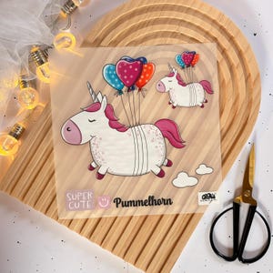 Könnte beinhalten: Ein weißes Einhorn mit rosa Mähne und Schweif fliegt mit einem Bündel bunter Ballons. Das Einhorn hält eine Schnur mit Ballons in den Farben Rosa, Rot, Blau und Weiß. Der Text "Super Cute" und "Pummelhorn" ist auf dem Bild gedruckt.