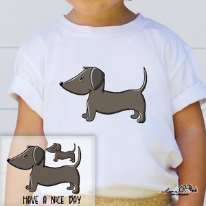 Könnte beinhalten: Weißes T-Shirt mit einem Cartoon-Bild eines braunen Dackels. Der Text "Have a Nice Day" ist unten auf dem Shirt aufgedruckt.