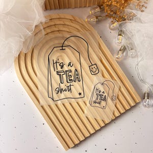 Könnte beinhalten: Zwei transparente Aufkleber in Form eines Teebeutels mit schwarzem Text, der "It's a TEA shirt" lautet, und einem Smiley. Die Aufkleber befinden sich auf einer Holzoberfläche.
