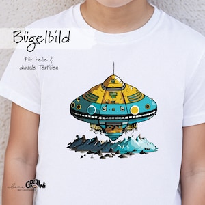 Könnte beinhalten: Ein weißes T-Shirt mit einem gelben und blauen Cartoon-UFO, das über einem Gebirge schwebt. Der Text "Bügelbild" und "Für helle & dunkle Textilien" ist auf dem Shirt aufgedruckt.