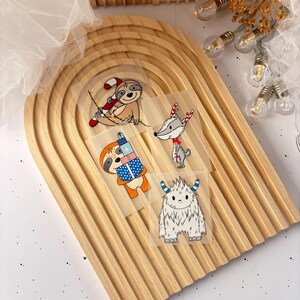 Könnte beinhalten: Eine Auswahl an weihnachtlichen Aufbüglern mit Cartoon-Charakteren. Die Designs umfassen Faultiere mit Zuckerstangen und Geschenken, ein Reh mit rotem Schal und einen Yeti mit blau gestreiften Hörnern. Ideal zum Aufbügeln auf Kleidung oder Accessoires.