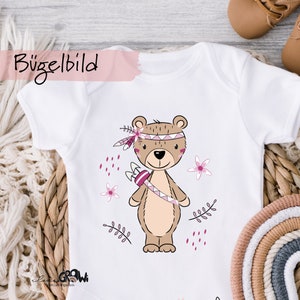 Könnte beinhalten: Weißer Baby-Body mit einem Cartoon-Bären, der einen Indianer-Kopfschmuck und einen Pfeil und Bogen trägt. Der Bär ist braun mit rosa und lila Akzenten. Der Text "Bügelbild" ist sichtbar.