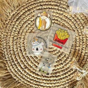 Könnte beinhalten: Vier illustrierte Sticker mit einem Affen, der eine Banane hält, einem Koala mit Kopfbedeckung, einem Wildschwein, das einen Roller fährt, und einer roten Schachtel Pommes Frites. Die Sticker haben deutschen Text, der "Kleiner Wilddecker", "Sonnenstrahlen" und "frittierte" lautet.