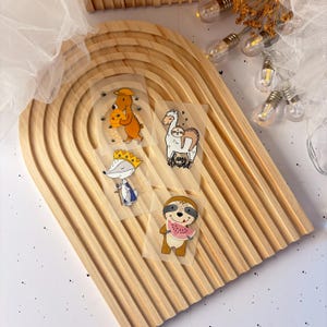 Könnte beinhalten: Auf einem hölzernen, bogenförmigen Tablett liegen vier transparente Aufkleber mit Cartoon-Tierdesigns. Die Aufkleber zeigen einen Bären mit Bienen, ein Lama mit dem Schriftzug "hang around", einen Fuchs mit Krone und ein Faultier mit Wassermelone.