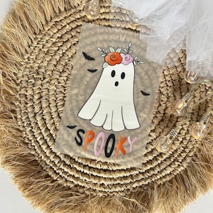 Könnte beinhalten: Ein Halloween-Design mit einem weißen Geist mit Blumenkranz und dem Wort "Spooky" in Orange und Pink. Schwarze Fledermäuse sind um den Geist herum verstreut. Das Design befindet sich auf einem hellbraunen Hintergrund.
