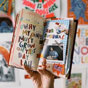 Puede incluir: Un diario abierto con páginas coloridas y texto escrito a mano que dice "¿Por qué mis granos deben crecer en este día lah..." El diario tiene una pegatina que dice "Diario de arte" y una foto de una persona sentada en un escritorio. El diario está sostenido en una mano con un tatuaje.