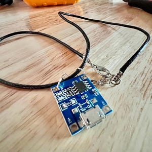May include: A blue electronic circuit board pendant with a black cord necklace. The pendant has silver connectors and a micro USB port. The necklace is on a light brown wooden surface.