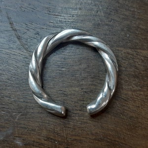 Puede incluir: Un brazalete de plata trenzado. El brazalete está hecho de una sola pieza de metal que está trenzada en un diseño similar a una cuerda.