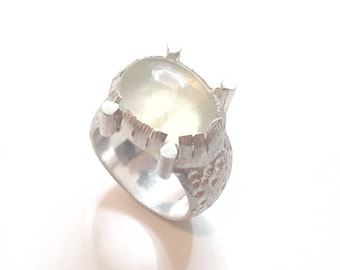 Puede incluir: Un anillo de plata con una banda texturizada y una gran piedra blanca de forma ovalada.