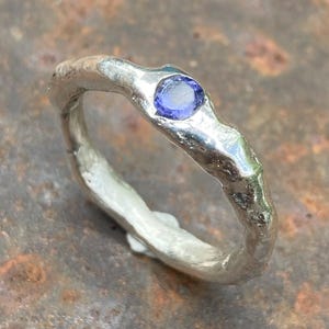 Op de afbeelding: Een zilveren ring met een getextureerde band en een kleine, ronde, paarse edelsteen.