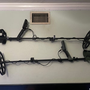 Può includere: Due metal detector neri sono montati su una parete grigio chiaro. I detector sono appesi orizzontalmente con le bobine di ricerca rivolte verso il basso. I detector sono collegati a cavi che sono collegati a prese di corrente.