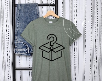 May include: A green t-shirt with a black graphic of a box with a question mark on top. The shirt is hanging on a wooden hanger against a white background with a black and white pattern.