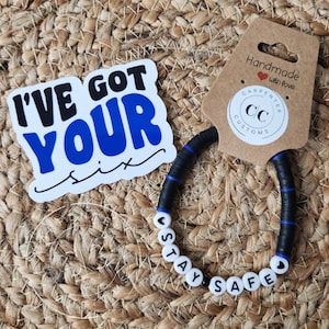 May include: A black and blue beaded bracelet with white beads that spell out "Stay Safe". The bracelet is handmade and has a small heart charm. The bracelet is accompanied by a sticker that says "I've got your six".