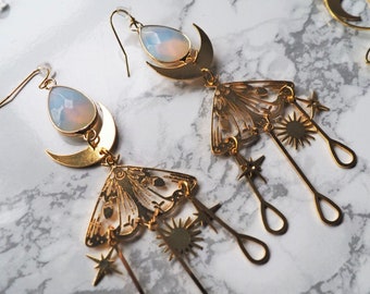 Puede incluir: Pendientes de tono dorado con una luna creciente y una piedra similar a la ópalo, un diseño de ala de polilla y colgantes de estrellas y soles.