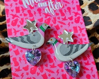 May include: A pair of silver star and purple heart dangle earrings featuring a gray dove with green accents. The earrings are displayed on a pink background with a white pattern and the text "Mallory Ever After".