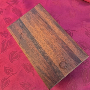 Peut inclure: Une boîte rectangulaire en bois avec un couvercle à charnière. La boîte est faite de bois veiné brun foncé et brun clair. Une petite indentation circulaire sert de poignée à l'avant. La boîte est sur une surface rouge.