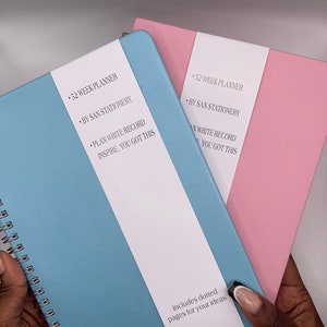 May include: Two 52-week planners, one blue and one pink, with the text "52 Week Planner", "By San Stationery", "Plan Write Record Inspire, You Got This", and "Includes dotted pages for your ideas".
