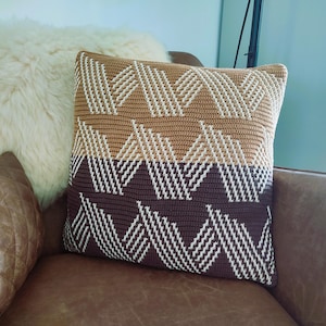 Op de afbeelding: Een vierkant decoratief kussen met een geometrisch patroon in bruin- en wittinten. Het kussen is gehaakt en heeft een getextureerde uitstraling. Het ligt op een bruine leren bank, met een fluffy wit kussen op de achtergrond.