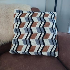 Op de afbeelding: Een vierkant decoratief kussen met een geometrisch patroon in wit, zwart, grijs en bruin. Het kussen ligt op een bruine leren stoel, met een fluffy, crèmekleurig kussen op de achtergrond.