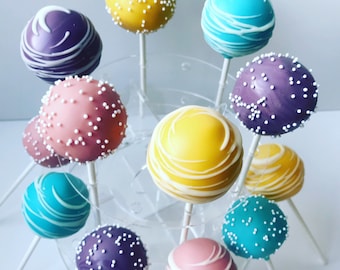 May include: A collection of 10 cake pops decorated with different colors and patterns. The cake pops are arranged on a clear plastic stand. The cake pops are decorated with white, pink, yellow, blue, and purple frosting. Some of the cake pops are decorated with white sprinkles.