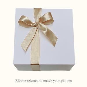 May include: A white gift box tied with a gold satin ribbon and bow. The ribbon wraps around the box and is tied in a decorative bow on top. The text at the bottom of the image reads: "Ribbon selected to match your gift box."