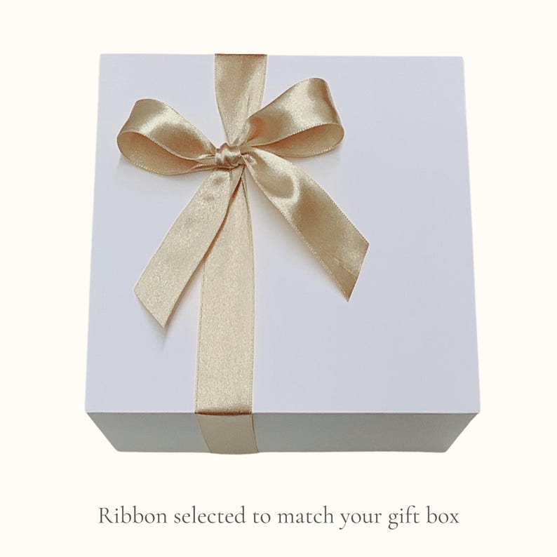 May include: A white gift box tied with a gold satin ribbon and bow. The ribbon wraps around the box and is tied in a decorative bow on top. The text at the bottom of the image reads: "Ribbon selected to match your gift box."