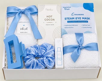 May include: A gift box containing a bag of Juniper & Eucalyptus bath salt with a blue ribbon, a box of Creamy White Hot Cocoa, an unscented Frshmorn steam eye mask, a blue hair claw clip, a blue satin scrunchie, a lip balm, and a white towel.