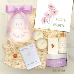 May include: A gift box containing a bag of lavender and eucalyptus bath salt with a purple ribbon, two white tea light candles, a box of matches, a small bottle of face mask, a Wildland super bar, and a jar labeled big hug with a floral card.