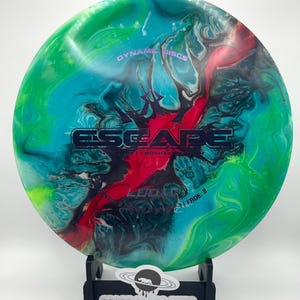 May include: A disc golf disc with a vibrant, marbled pattern in green, blue, and red. The disc is emblazoned with the word "ESCAPE" and the text "DYNAMIC DISCS". It is displayed on a black stand, with the text "SPACE RHINO DYES" visible on the stand.