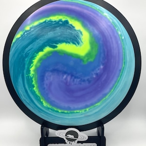 May include: A disc golf disc with a vibrant, swirling design in shades of blue, teal, purple, and neon green. The disc has a black rim and is displayed on a black stand. The text "SPACE RHINO DYES" is visible on the stand.