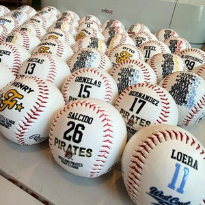 May include: A collection of white baseballs with red stitching, each customized with black and colored text including names, jersey numbers, and team logos such as Pirates, West Coast Sox, and Little League All-Stars 2026 Champions.