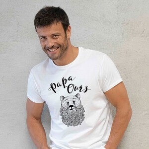 May include: White t-shirt featuring a black and white illustration of a bear with a beard and moustache. The text "Papa Ours" is written above the bear.