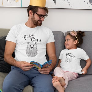 May include: A father and daughter wearing matching white t-shirts with a bear graphic and the text "Papa Ours" and "Petit Ours". The father is wearing a gold crown and holding a book. The daughter is wearing a pink headband. They are sitting on a grey sofa.
