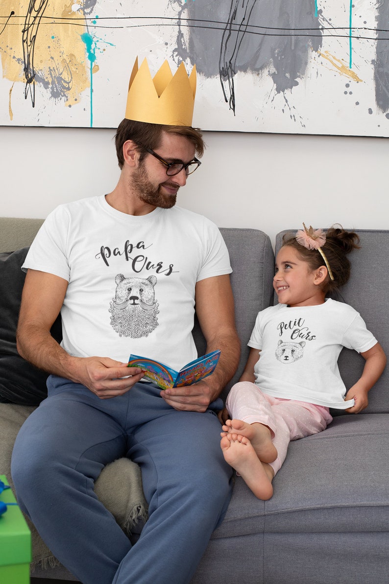 May include: A father and daughter wearing matching white t-shirts with a bear graphic and the text "Papa Ours" and "Petit Ours". The father is wearing a gold crown and holding a book. The daughter is wearing a pink headband. They are sitting on a grey sofa.