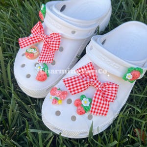 Puede incluir: Zuecos blancos decorados con lazos de cuadros rojos y adornos de fresas. Los adornos incluyen fresas, flores y una cesta. Los zuecos están sobre hierba verde.