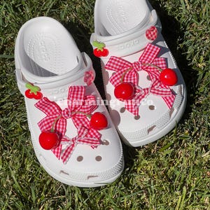 May include: A pair of white Crocs decorated with red gingham bows and bright red cherry charms. The shoes are set on a bed of green grass, showcasing the playful and summery design.
