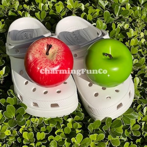 May include: White clogs with a red apple and a green apple on top. The clogs have a textured surface and a hole pattern.