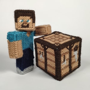 Könnte beinhalten: Eine braune und beige gehäkelte Minecraft-Figur, Steve, steht neben einer braunen und beige gehäkelten Truhe. Steve trägt ein blaues Hemd und eine dunkelblaue Hose.