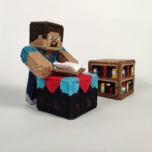 Könnte beinhalten: Eine Minecraft-Figur aus Garn, die ein blaues Hemd und eine braune Hose trägt, bastelt einen weißen Block auf einem Basteltisch. Der Tisch ist aus Garn und hat ein schwarzes, violettes, blaues und rotes Design. Ein Bücherregal aus Garn befindet sich im Hintergrund.