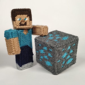 May include: A blue and brown yarn craft of a Minecraft character standing next to a gray and blue yarn block.