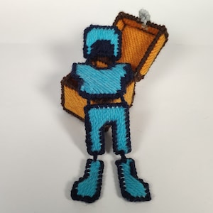 May include: A handmade, pixelated figure of a person in blue armor, holding an open treasure chest. The chest is orange with brown trim. The figure is made of yarn and has a retro video game aesthetic.