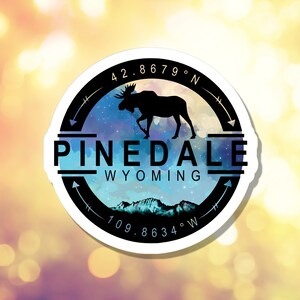 Puede incluir: Una pegatina redonda con un diseño en blanco y negro. La pegatina presenta la silueta de un alce sobre un fondo de acuarela azul y morado con estrellas. El texto "PINEDALE WYOMING" está impreso en blanco sobre una banda negra. Las coordenadas "42.8679° N 109.8634° W" están impresas en blanco sobre una banda negra.