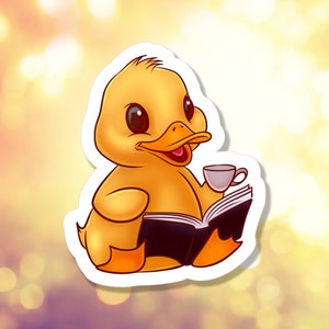 Puede incluir: Un pequeño pato amarillo con una expresión alegre está sentado con un libro y una taza de té. El patito está sosteniendo el libro con su ala derecha y la taza con su ala izquierda. El libro es negro con una cubierta blanca.