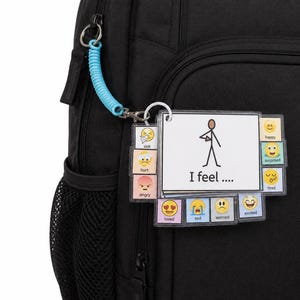 May include: A black backpack featuring a communication keychain with a light blue coil. The keychain displays a central card with a stick figure and the text I feel, surrounded by smaller cards showing various emoji faces labelled sick, hurt, angry, loved, sad, worried, excited, tired, surprised, and happy.