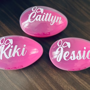May include: Three pink plastic Easter eggs with bunny ears and the names Caitlyn, Kiki, and Jessica written in white.