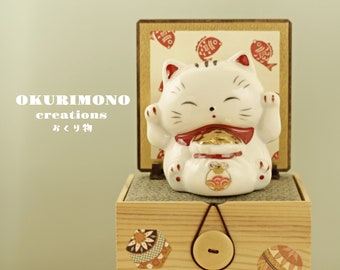 May include: A white ceramic Maneki-Neko, or lucky cat, figurine with raised paw, sitting in a wooden box with a gold coin in its paw. The box has a red and gold design with fish patterns. The text "OKURIMONO creations おくり物" is visible on the box.