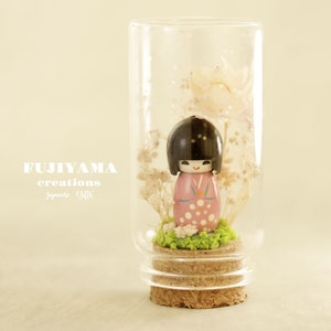 Könnte beinhalten: Eine kleine, rosa-weiße japanische Puppe in einem traditionellen Kimono ist in einem Glasgefäß mit getrockneten Blumen und Moos ausgestellt. Das Gefäß hat einen Korkverschluss und ist mit "FUJIYAMA creations Japanese Gifts" beschriftet.