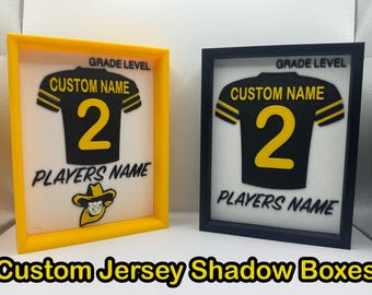 May include: Two framed shadow boxes with a football jersey design. One frame is yellow, the other black. Each jersey is black with yellow accents and the number 2. Text includes "GRADE LEVEL", "CUSTOM NAME", and "PLAYERS NAME". The bottom text reads "Custom Jersey Shadow Boxes".